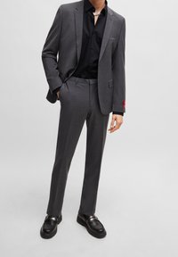 Completo grigio con vestibilità sartoriale, camicia nera e scarpe nere. Accento rosso sul polsino. Tessuto morbido con un design formale.