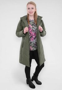 Manteau long matelassé vert olive avec capuche, doté de poches latérales zippées. Associé à un haut à motif floral et des leggings noirs. Bottes noires à la cheville.