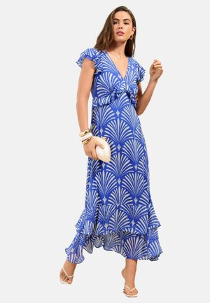 Kvinde iført en blå og hvid mønstret kjole med flæser, holder en hvid clutch og er accessoriseret med gyldne armbånd og øreringe i ringform.