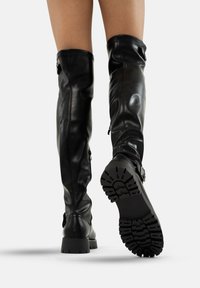 Piernas con botas altas de cuero negras hasta la rodilla, con suelas gruesas y estriadas, una bota levantada mostrando el detallado patrón de la suela.