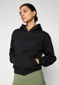 Black zip-up hoodie with a large hood, front pockets, and ribbed cuffs. Made from a soft fabric, paired with olive green bottoms.