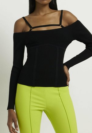 Femme portant un top côtelé noir à épaules dénudées avec de fines bretelles et un pantalon ajusté jaune vif, debout avec une main sur la hanche.