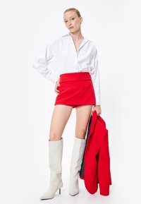 Hvid skjorte med knapper, rød mini skirt og knæhøje lysegrå støvler. Model holder en rød blazer og står foran en ensfarvet hvid baggrund.