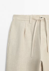 Light beige linen pants with front pleats, belt loops, and a matching fabric tie belt at the waistband.