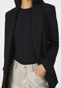 Henkilö, jolla on yllä musta bleiseri mustan paidan päällä ja vaaleanharmaat krokotiilinkuosiset housut, käsi taskussa, kuvattu kaulasta keskivartaloon.