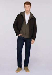 Svart jacka med huva över en mörkgrön huvtröja med dragkedja, kombinerad med mörka jeans och beige sneakers. Enkel design utan framträdande mönster.