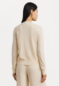Pulover de culoare crem, cu mâneci lungi, cu mansete și tiv cu model în relief. Textură netedă, design simplu, fără modele vizibile sau detalii din metal.