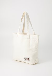 Kreminės spalvos drobės rankinė su dviem audinio rankenomis. Apatinėje kairėje kampe yra ruda logotipo etiketė. Paprasta, stačiakampė forma.