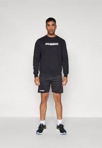 Sort langærmet sweatshirt med "HUMMEL" logo, parret med sorte shorts og atletiske sko med accenter i grøn og blå.