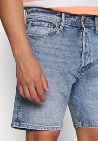 Pantalones cortos de denim azul claro con un corte holgado, diseño de cinco bolsillos y costuras visibles. Textura suave y ligero desgaste en la tela.