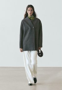 Cappotto doppiopetto in lana grigia con bottoni d'argento, abbinato a pantaloni bianchi e borsa nera. Sciarpa verde che aggiunge un dettaglio a contrasto.