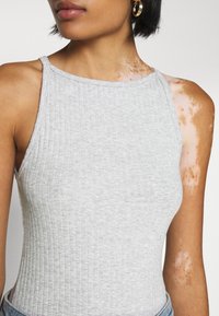 Canottiera grigia a costine con collo alto e spalline sottili, caratterizzata da una silhouette aderente e una texture sottile. Variazioni di tono della pelle visibili.