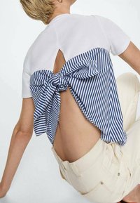 Top blanco de manga corta con la parte posterior a rayas azul y blanca que presenta un gran lazo. Tejido ligero y corte holgado, combinado con pantalones cortos beige claro.