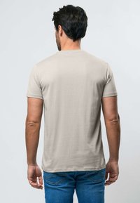 Muškarac s tamnom kosom, obučen u jednostavnu bež majicu kratkih rukava i plave traperice, stoji okrenut leđima prema svijetloj pozadini.