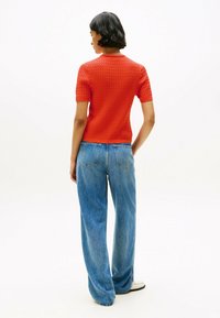 Rotes kurzärmliges Stricktop mit einem strukturierten Muster, kombiniert mit hellblauen weiten Jeans und weißen Schuhen. Ansicht von hinten.