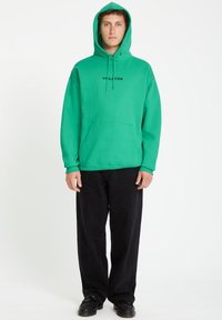 Groene hoodie met trekkoorden en een voorzak, met het zwarte "Volcom" logo. Gecombineerd met zwarte broek met wijde pijpen en zwarte schoenen.