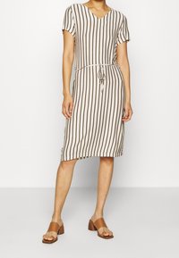 Robe rayée en beige et marron, décolleté en V, manches courtes, taille à cordon, longueur genou, tissu doux, associée à des sandales à talons marron.