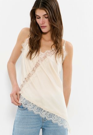 Femme portant un caraco crème orné de dentelle diagonale et un jean bleu clair, regardant vers le bas avec une main dans la poche.