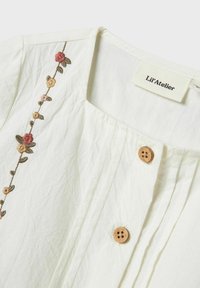 Blusa bianca con ricamo floreale sulla parte frontale, caratterizzata da bottoni in legno e texture plissettata vicino al collo. Etichetta: Lil'Atelier.