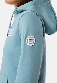 Lichte blauwe hoodie met een kangoeroezak, textuurstof en een ronde logopatch op de linker mouw.