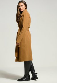 Brun uldfrakke med bælte, store kraver og lange ærmer, båret med knæhøje sorte læderstøvler med sidestrap detajle.