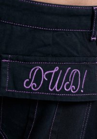 Tissu noir avec des coutures violettes et des lettres brodées "DW!" sur une poche ou un rabat, montrant un travail de couture détaillé.