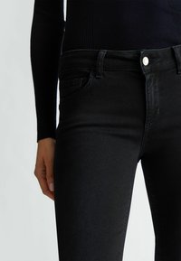 Jean en denim noir avec une coupe slim, dotés de passants de ceinture, de poches avant et d'un détail de bouton en métal argenté à la taille. Texture lisse.