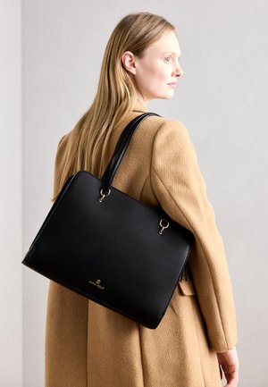 Mujer con cabello largo y rubio que lleva un abrigo camel y lleva un gran bolso de cuero negro con detalles dorados en el hombro.
