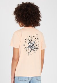 Korte mouwen, perzikkleurig t-shirt van zachte stof, met een zwart spinnenafbeelding en webontwerp op de achterkant met decoratieve accenten.