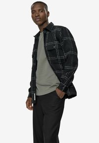 Sort rutet shacket i blødt stof, med hvide gitterlinjer, frontlommer og knaplukninger, parret med en grå crew-neck t-shirt og sorte bukser.