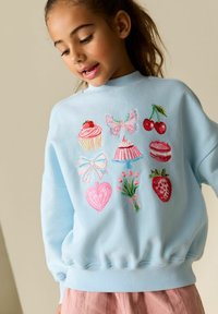 Sweat-shirt bleu clair avec des motifs brodés incluant des cupcakes, des cerises, un papillon et des fleurs. Texture douce et coupe confortable.