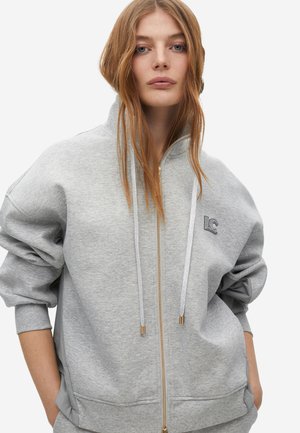 Sudadera con capucha gris con cremallera hecha de material suave, con cuello alto, puños acanalados, bolsillos delanteros y un pequeño logotipo en el pecho.