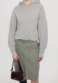 Persona vistiendo una sudadera gris claro y una falda verde salvia, sosteniendo un pequeño bolso marrón oscuro con una correa corta en una mano.