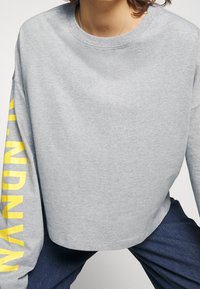 Gray cropped sweatshirt with a round neckline and long sleeves, featuring bold yellow text along the left sleeve. Soft fabric texture.