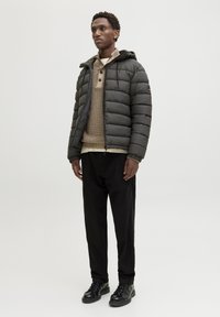 Padded czarna kurtka z kapturem, z zapięciem na zamek błyskawiczny z przodu i elastycznymi mankietami, noszona na beżowym swetrze oraz czarnych spodniach, w połączeniu z czarnymi butami.