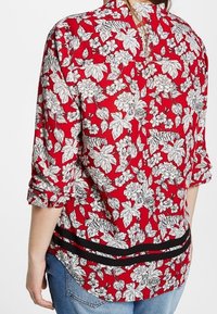Blusa floreale rossa con motivi botanici in bianco e nero, orlo curvo e una striscia nera in vita. Maniche lunghe arrotolate.