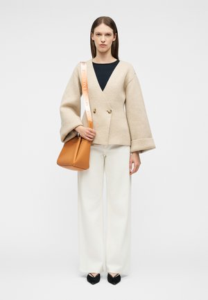 Γυναίκα που φοράει μπεζ ζακέτα με διπλό κουμπί, λευκό παντελόνι με φαρδιά μπατζάκια, μαύρες γόβες και κρατά μια καφέ τσάντα ώμου με φαρδιά λουράκι με λογότυπο.