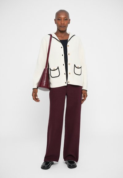 Cárdigan blanco de canalé con ribete negro y bolsillos, combinado con pantalones de pierna ancha burdeos y zapatos negros. La modelo sostiene un bolso burdeos.