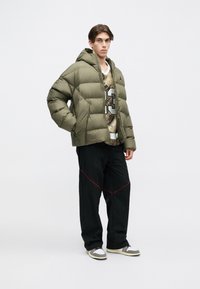 Young man wearing an olive green puffer jacket, camo shirt with number 23, loose black pants with red stitching, and gray-white sneakers.