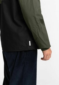 Windbreaker-Jacke mit schwarzem Körper und olivgrünen Ärmeln. Leichtes Material, glatte Textur, elastische Bündchen, ausgestattet mit einem kleinen Markenetikett.