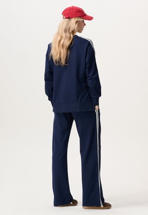 Marineblauw trainingspak met contrasterende witte zijstrepen, voorzien van een losse pasvorm en zachte stof. Gecomplementeerd met een rode pet en bruine schoenen.