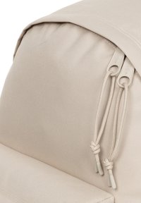 Mochila de tela color beige claro con superficie texturizada, cierres dobles con cremallera y tiradores de cuerda, y una forma ligeramente redondeada en la parte superior.