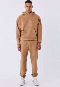 Tan fleece hoodie met een kangoeroezak en gouden logo, gecombineerd met bijpassende taps toelopende broek en witte sportschoenen. Glad materiaal.