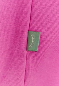 Fuchsiová pletená tkanina s texturovaným povrchem, na boku malá zelená visačka s bílým designem řas.