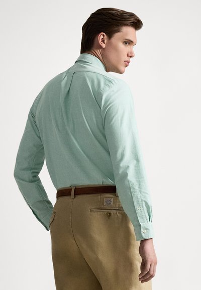 Šviesiai žalios spalvos marškiniai su sagomis ir ilgomis rankovėmis, su nugara siūtu įsiuvu ir apvaliu apačios kirpimu, derinami su rudos spalvos kelnėmis ir rudai diržu.