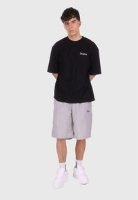 Czarny bawełniany t-shirt z logo "Dropsize", w połączeniu z jasnoszarymi spodniami w kratę. Widać białe sneakersy i skarpetki. Proste tło.
