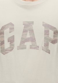 Chemise beige clair avec les grandes lettres G, A et P dans un motif camouflage discret sur la poitrine.