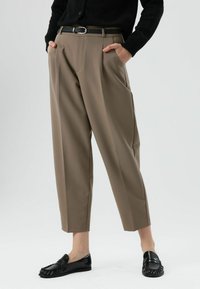 Pantalon beige à coupe ajustée, avec plis à l'avant, poches et ceinture. Associé à des mocassins noirs, sur un fond clair.