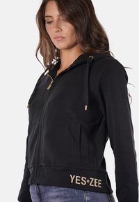 Donna che indossa una felpa nera con zip e coulisse, con la scritta "YES•ZEE" ricamata in oro vicino all'orlo, abbinata a jeans blu.
