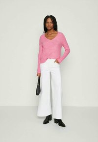 Moteris su ilgais kaspinais, dėvinti rožinį V formos kaklo megztinį, baltas plačias kelnes, juodus kulkšnies aulinukus ir laikanti mažą juodą rankinę.